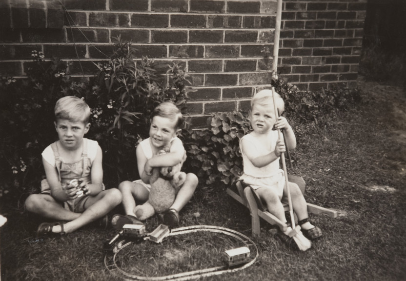 Three boys playing with a toy train set, teddy bear and other toys on the lawn of a backyard.Three brothers Darryl, John and Trevor Hawksworth, in the backyard of the Hawksworth family home, Strathmore, 1949.