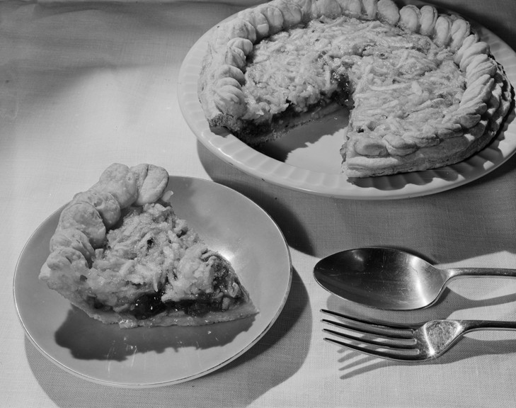 Product photograph of a pie dessert, a slice has been removed and placed on a plate in front of it. There is a fork and spoon placed next to it.