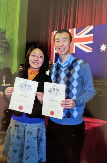 A couple standing in front of the Australian flag holding certificates