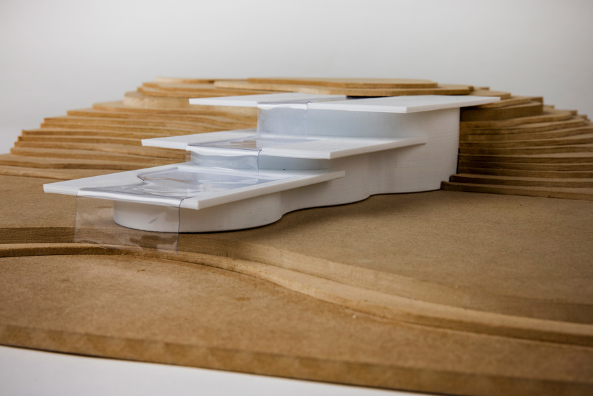 A close-up of an architectural model. A white building is set into a hilly landscape, inferred by topographical lines. The building has three stepped levels connected by a man-made waterfall.