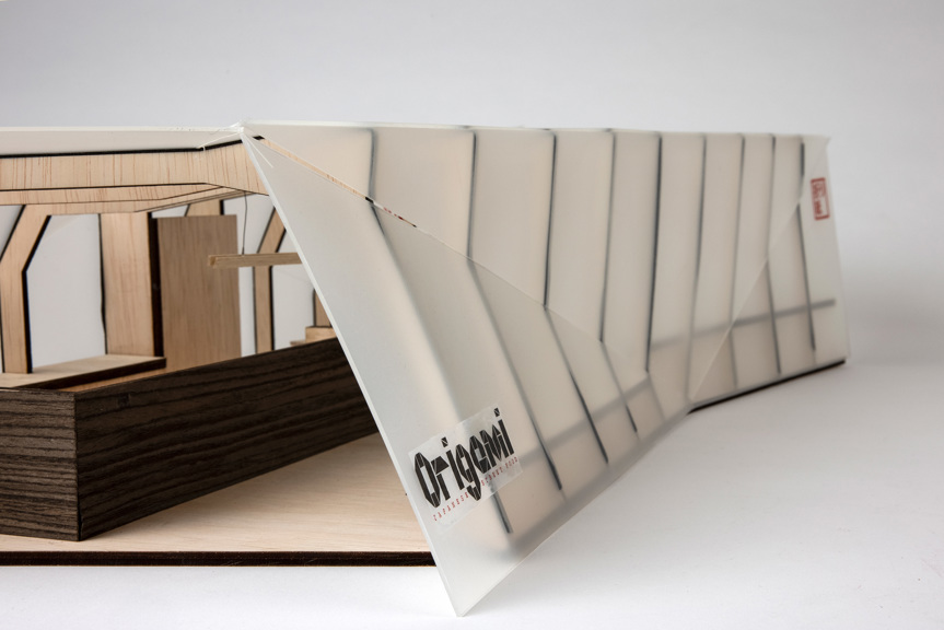 A photo of an architectural model. Angular forms are used to convey the aesthetic of origami. Dark and light woods are used in the interior. A red logo in Japanese characters is stamped on the outside.