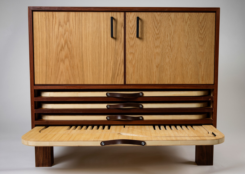 The image shows the chair packed down into a cabinet form. It is made of a light coloured wood, with darker wood edging. It has leather handles to help with assembly and disassembly.