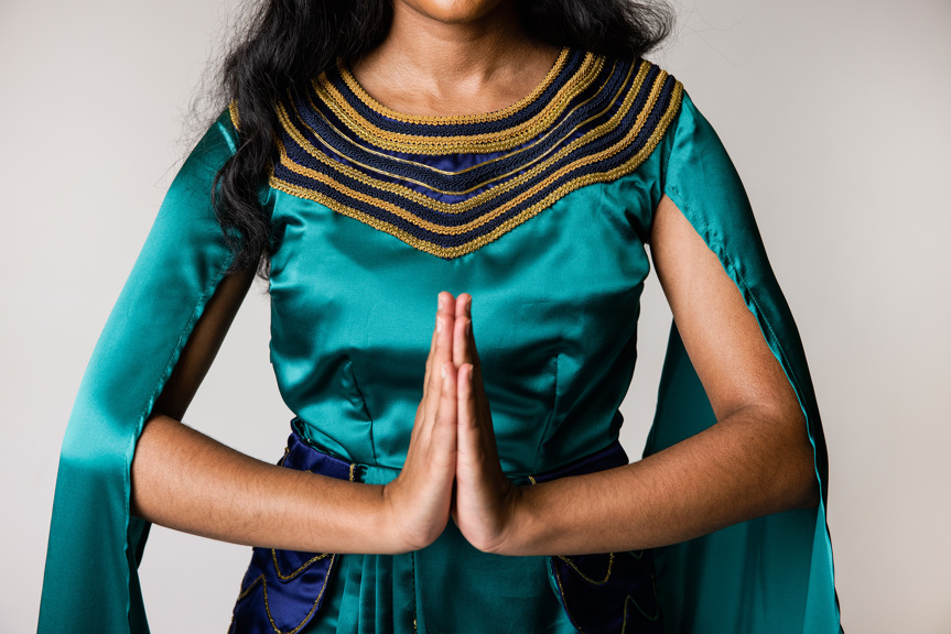 A person with long, dark hair is shown wearing a garment made from emerald green silk. The garment has gold and navy blue embellishments around the neckline. The wearer is holding their hands together as if in prayer.