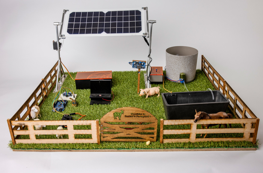 An image of a model of a farm. It is surrounded by a fence which contains a solar panel, a water tank and a water trough, and toy farm animals.