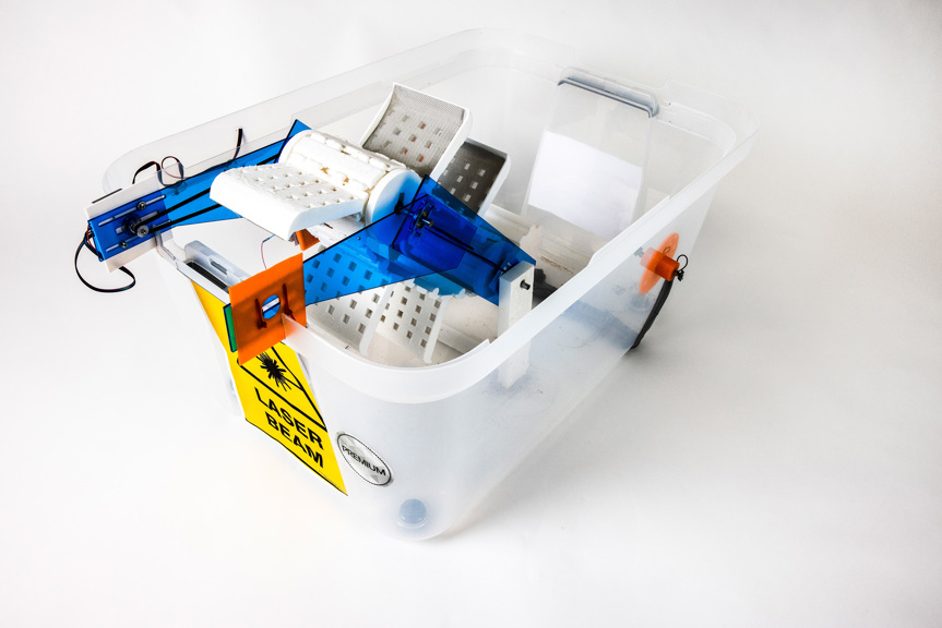 A clear plastic tub has a yellow label reading “laser beam”. Inside the tub are various motorized components, most notably a conveyer belt system. Blue and orange acrylic arms secure the belt.