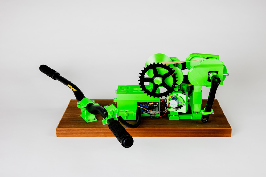 An image of a prototype transmission system. It is fluro green and fixed onto a panel of wood. Handle bars are placed at the front to mimic a bike.