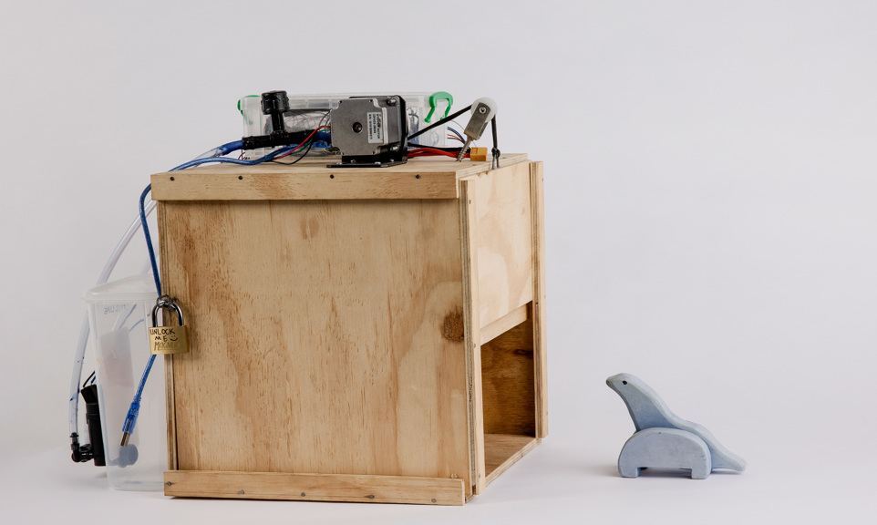 An image of the tracking device. It is a wooden box with a trapdoor that is activated by a sensor. A water container is fixed to the rear. A wooden toy is placed in front.