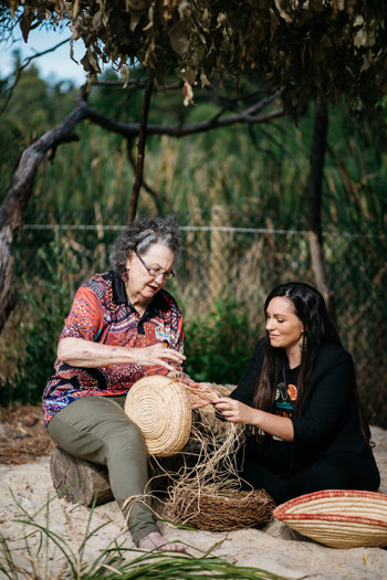 An older woman on the left is sitting on a log with a younger woman sitting in the sand weaving baskets from natural fibress