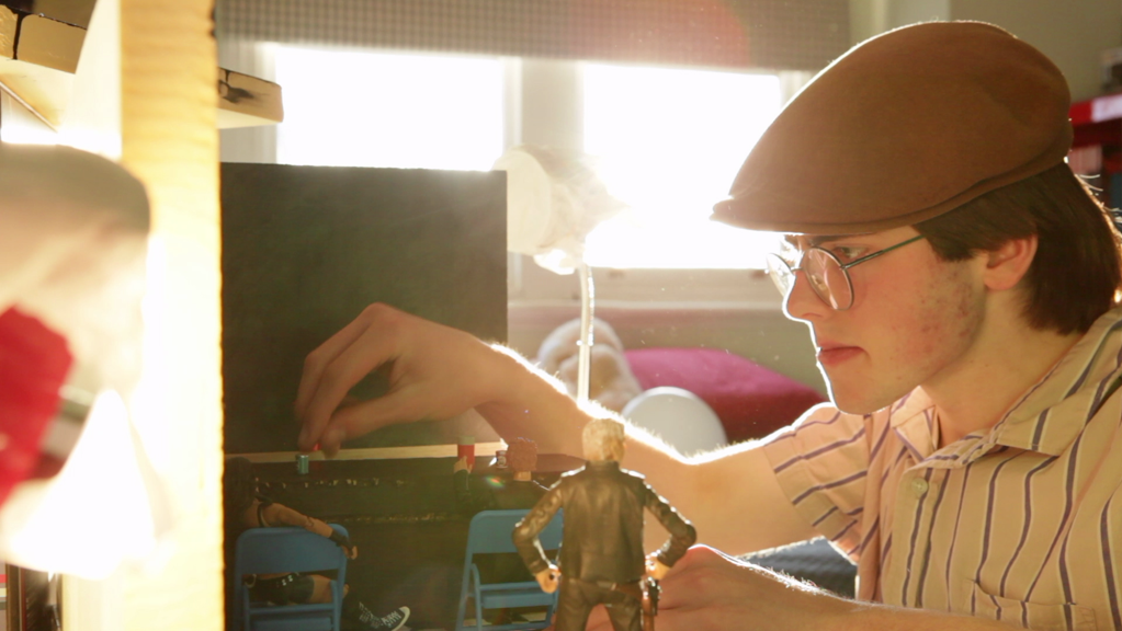 The still from ‘Magnum Opus’ shows a man wearing a flat cap, glasses, and a striped shirt. He is positioning some figurines for a stop motion shoot.