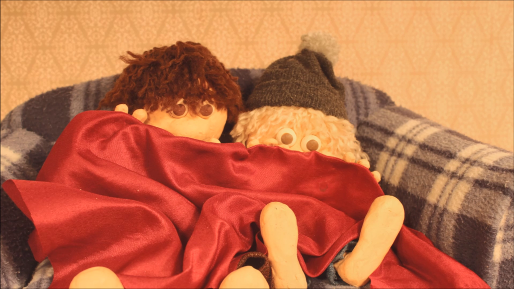 The still from the film ‘Quarantined’ shows two Claymation figures. The figures are peering out from underneath a red blanket with their feet poking out.