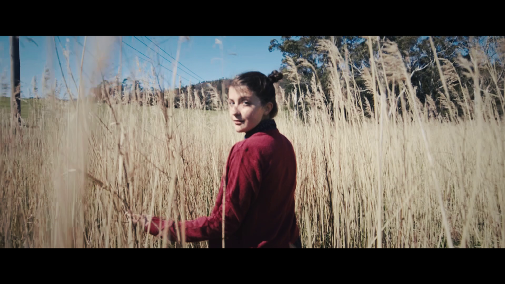 The still shows a woman standing in a field of wheat. She is turning to face the camera and has a neutral expression. There are trees and a blue sky in the background.