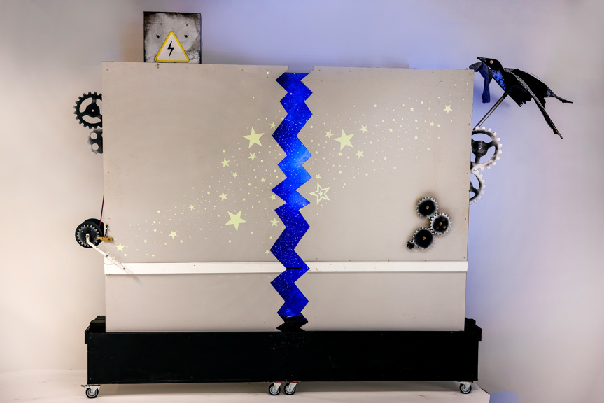 A photo of a grey rectangular wall on wheels. Two sides pull apart to reveal a blue galaxy. A bird holding a sock protrudes from the left-hand side.