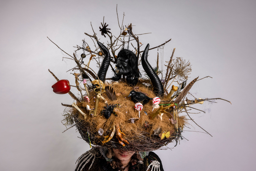 A photograph of a headpiece made to look like a nest. It is decorated with spindles, necklaces, bones, feathers, twigs, spiders, lollipops and chicken feet. A black voodoo head sits between two horns.