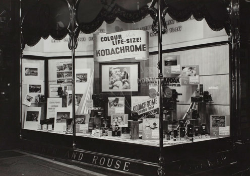 A shop front display case. Cameras and accessories on floor of case, plus posters and a large sign promoting Kodachrome colour film.