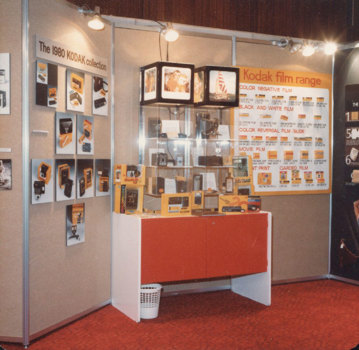 Tiered carpet board panels with photographs and display panels mounted on them. Red and white table with Perspex display cases above filled with products and labels. Cubed light boxes at top of display.