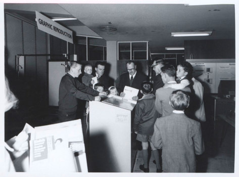 Group of men and young boys gathered around a piece of equipment in an exhibition display stand. 