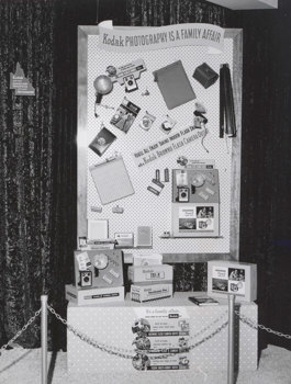 Exhibition display panel. The panel, set against a crushed velvet curtain, has products and signage suspended from it. A chain link barrier fence is visible in the foreground.