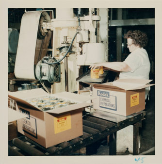 Woman in white overcoat putting tins from sealing machine into boxes of sealed products on conveyor belt in foreground.