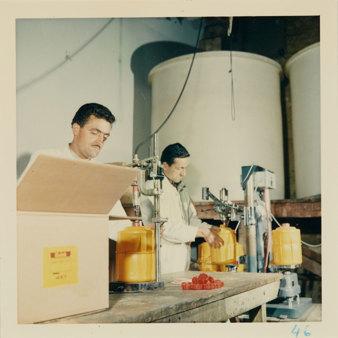 Two male workers in white overalls with yellow plastic containers underneath hoses and taps. Two large vats in background and box in foreground.
