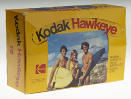 Yellow camera box with a photograph of two men and woman wearing swimwear outdoors. The two men are holding surf boards. Black and read text "Kodak Hawkeye"
