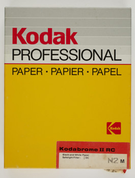 Rectangular box with yellow cardboard lid and black cardboard base. A sticker in lower right corner is printed with product details and seals the box.