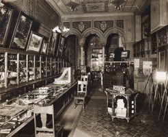 A shop interior, display counters and cases. Photographs hanging on walls. Chairs for clients to be served in. There are columns at the rear of the store with elaborate paint and plasterwork above them. The letter 'K' is painted in the centre of wall and ceiling friezes.