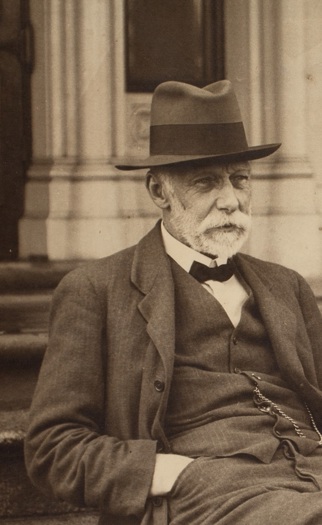 A bearded, seated man in a hat and bow tie