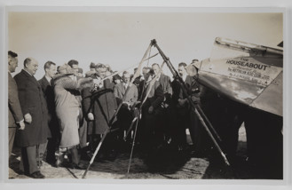 Crowd of men and women gathered at back of plane, with woman stretching out a large slingshot device. The plane has writing on the side reading: 'THE/ ROUSEABOUT / Presented to / The AUSTRALIAN AERO CLUB N.S.W / SECTION / By J.J. ROUSE ESQ.'