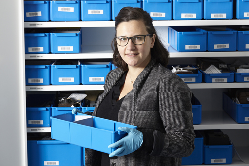 Woman in jacket and blue gloves holding blue plastic tote with a cupboard of totes behind her