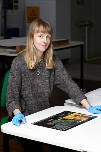 Woman sitting at table with poster in front of her. She is wearing blue gloves and a grey jumper.