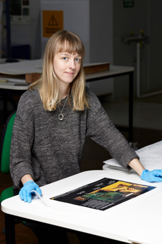 Woman sitting at table with poster in front of her. She is wearing blue gloves and a grey jumper.