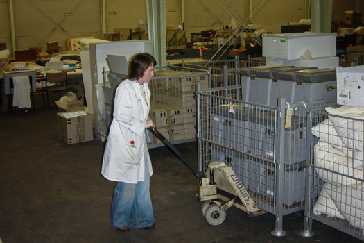 Woman in jeans and white laboratory coat pushing pallet of plastic crates in a warehouse
