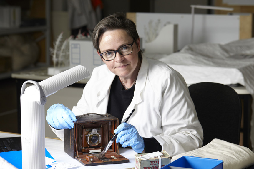 Woman in white laboratory coat seated at a table, wearing blue gloves and brushing a wood, leather and brass camera