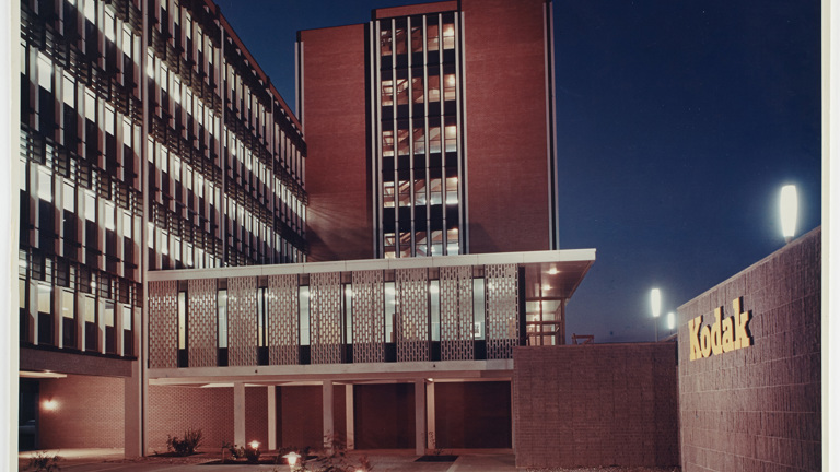 Building 8 in the newly constructed Kodak Australasia Pty Ltd factory complex in Coburg, circa 1965. Exterior view of a multi-storey building at night, with two sections of the building converging in an 'L' shape. Left section of the building has fewer storeys. At ground level there is an underground car park area. To the right is a low lying wall with "Kodak" signage. In the foreground is paved and grassed areas.