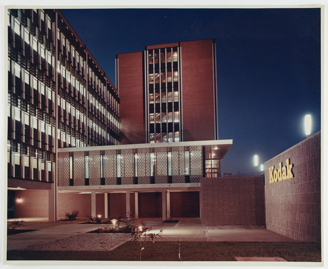 Building 8 in the newly constructed Kodak Australasia Pty Ltd factory complex in Coburg, circa 1965. Exterior view of a multi-storey building at night, with two sections of the building converging in an 'L' shape. Left section of the building has fewer storeys. At ground level there is an underground car park area. To the right is a low lying wall with "Kodak" signage. In the foreground is paved and grassed areas.