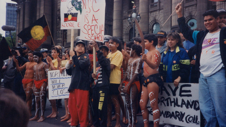 Group of people standing on the steps of parliament house holding placards and aboriginal flags