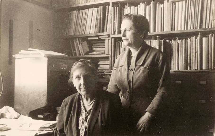 A black and white photo of two women, one seated, in front of a bookcase