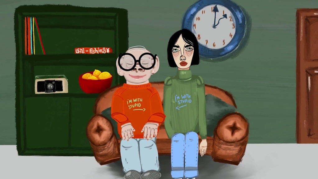 A still from an animated film, showing two people sitting together on a couch. They wear matching jumpers that read ‘I’m With Stupid’, pointing to one another. A wall clock and bookshelf are in the background.