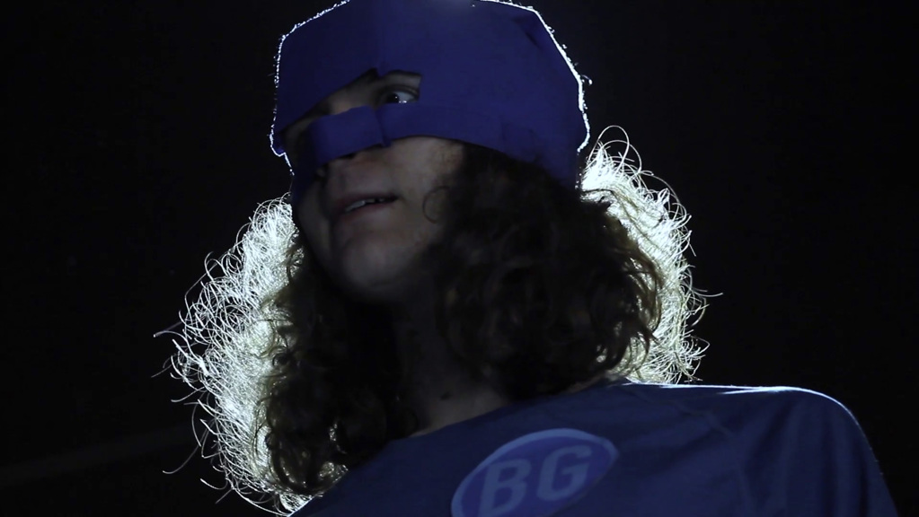 A young man with long hair wears a blue superhero mask and matching top. A cool light highlights his hair from behind and he has a surprised expression on his face. 