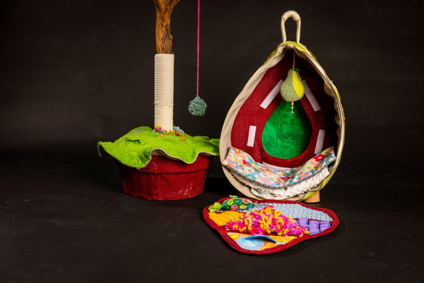 A cat carrier and scratching post next to each other. The cat carrier is shown open; it has a felt pear hanging from the top, and the inner wall is covered in different textured materials.