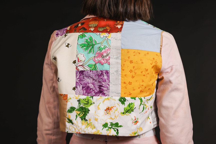 The back of a long-sleeved jacket is shown, with various colourful patterned panels, including florals, bees, and farm animals. The sleeves of the jacket are a light pink.