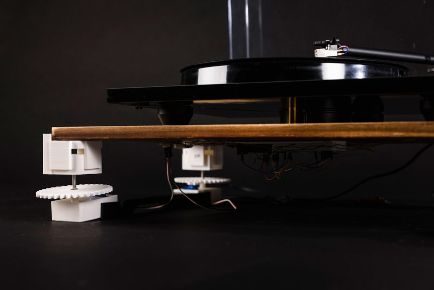 A close-up photograph shows a record player floating above a wooden shelf. White leveller components are installed on the corners of the shelf, and wires are visible underneath.