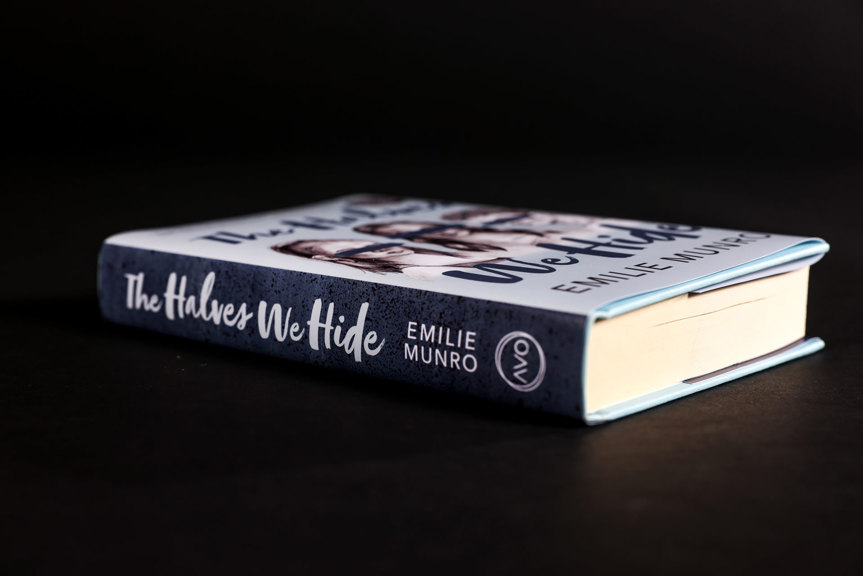 A blue book is photographed lying face up. The book’s spine reads “The Halves We Hide, Emilie Munro”. The Avo logo appears at the base of the spine.