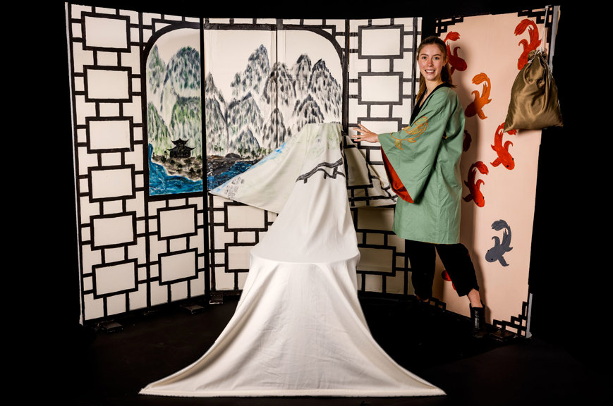 A painted wall features a window and wallpaper of goldfish. Through the window is a landscape of hills, water and a temple. The designer models the costume, a green Yi with wide sleeves and embroidered goldfish, and demonstrates the transformation of the set to represent a ghost.