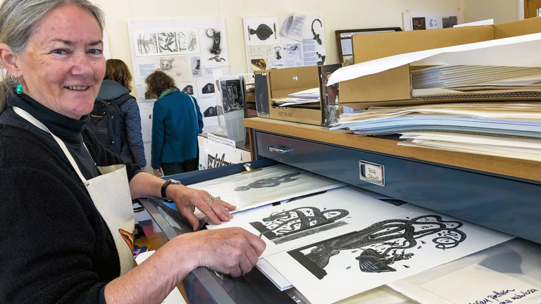 a photograph of a silver haired woman wearing an apron in front of a large drawer filled with black and white artistic prints