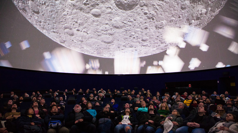 dozens of people reclining in chairs looking up at a giant projection of the moon 