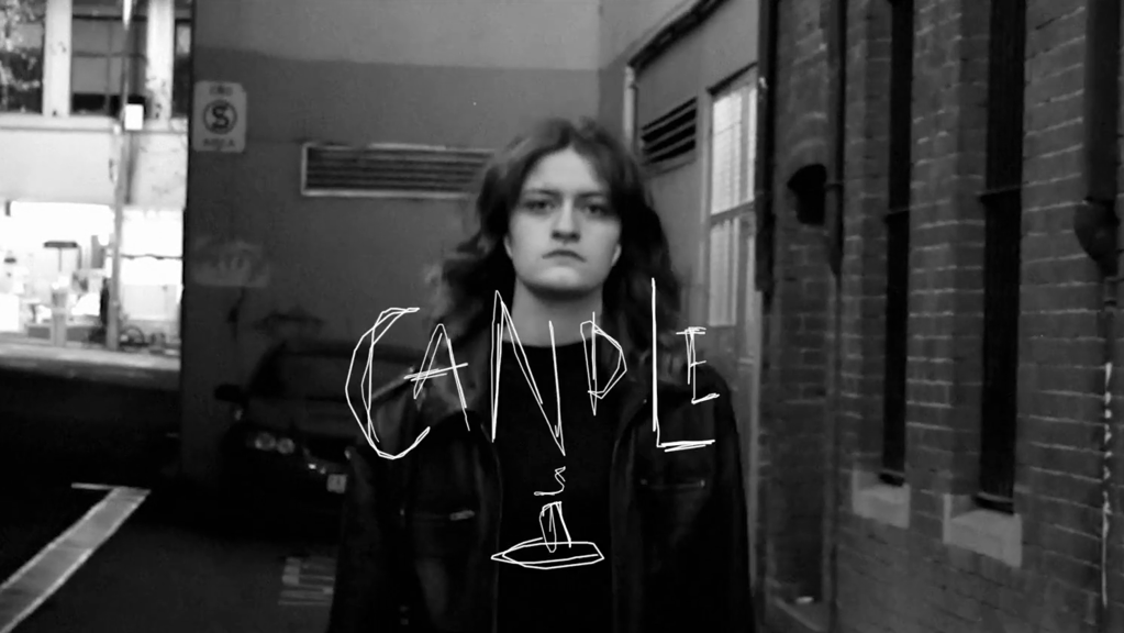 A film title screen in black and white. A young woman walks down an alleyway facing the camera. White text is overlaid reading ‘Candle’, with an illustration of a candle underneath.