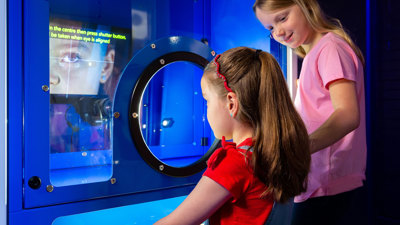 A child stands at an interactive blue exhibit featuring a circular viewing device, while an child in a pink shirt stands nearby. A screen in the background displays illuminated instructional text and a large close-up image of an eye. The scene is lit with bright blue light. 