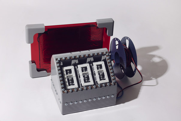 Grey, 3D printed rectangular base unit featuring three mechanical number displays, the entire top panel surrounded by a rectangle of visible screws. Behind it sits a red, transparent rectangular component and next to it is a plastic reel of wiring.