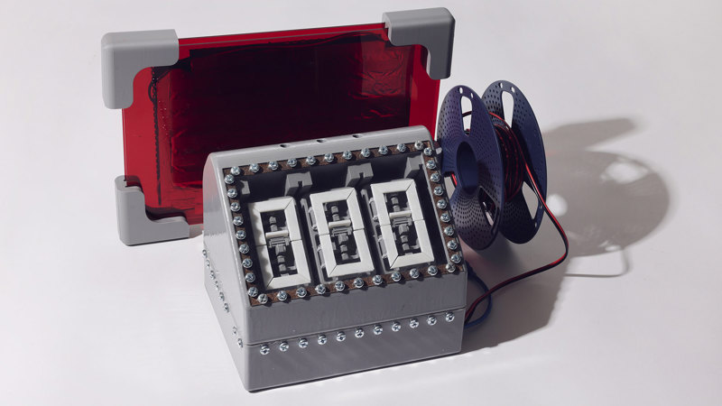 Grey, 3D printed rectangular base unit featuring three mechanical number displays, the entire top panel surrounded by a rectangle of visible screws. Behind it sits a red, transparent rectangular component and next to it is a plastic reel of wiring.
